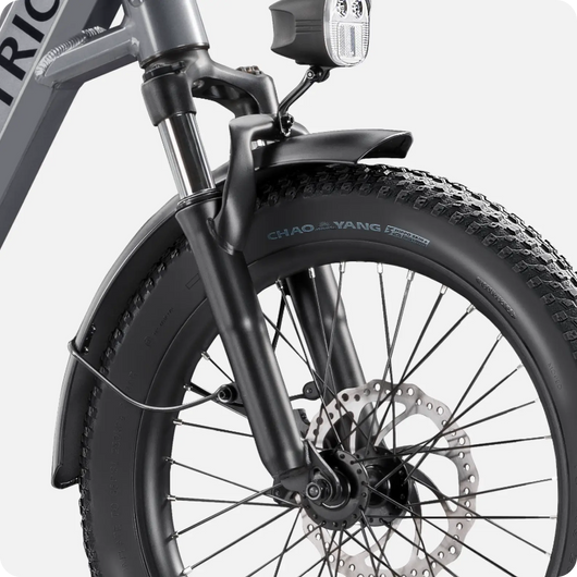 Close-up view of the Velotric Fold 1 electric bicycle's front wheel, fork, and headlight, featuring disc brakes and a thick treaded tire.