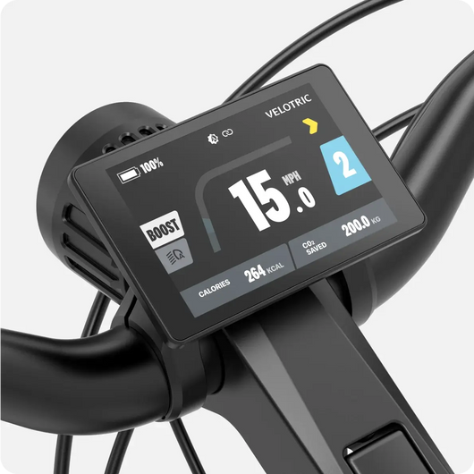 Close-up of a digital display on a Velotric - Discover 2 handlebar showing speed (15 mph), battery level (100%), mode (2), calories burned (264 kcal), and CO2 saved (20.0 kg). Velotric logo on screen.
