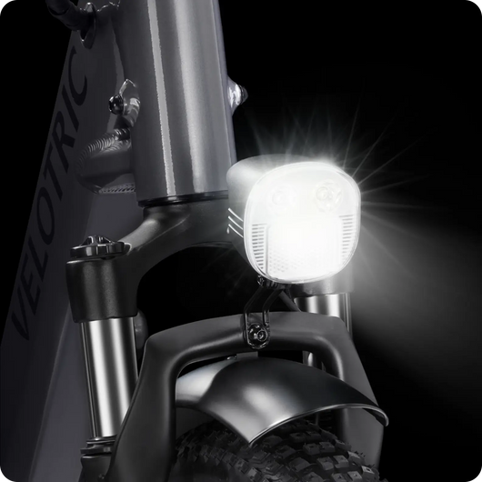 Close-up of a Velotric - Fold 1 eBike's headlight turned on, illuminating from the front of the bike, with the brand "Velotric" visible on the frame.