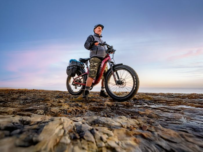 At sunset, a person in camo shorts and a jacket stands on rocky terrain with the Velotric - Nomad 2 all-terrain eBike, framed by a clear sky.