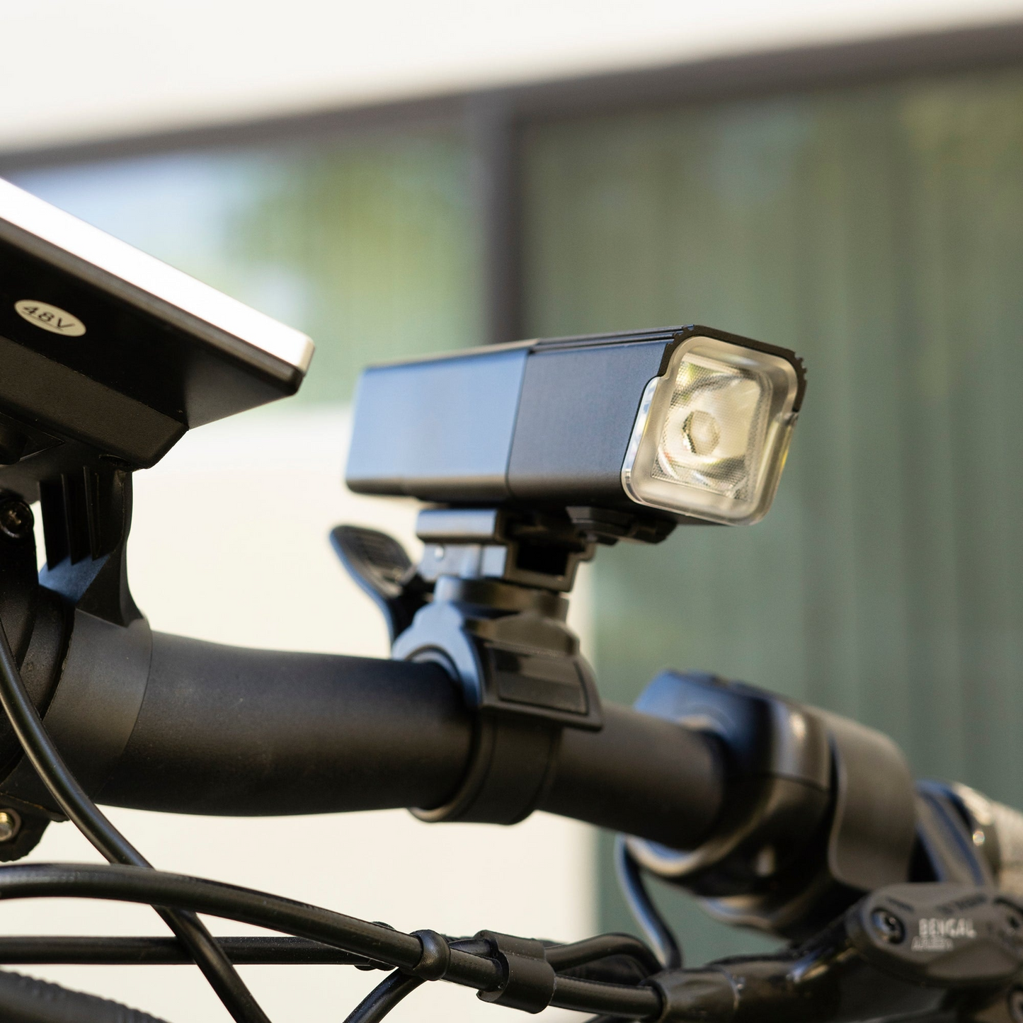 A close-up of a black bicycle handlebar shows the Aventon V10-500 headlight, which is USB rechargeable and features an adjacent bell, highlighting excellent bike compatibility.