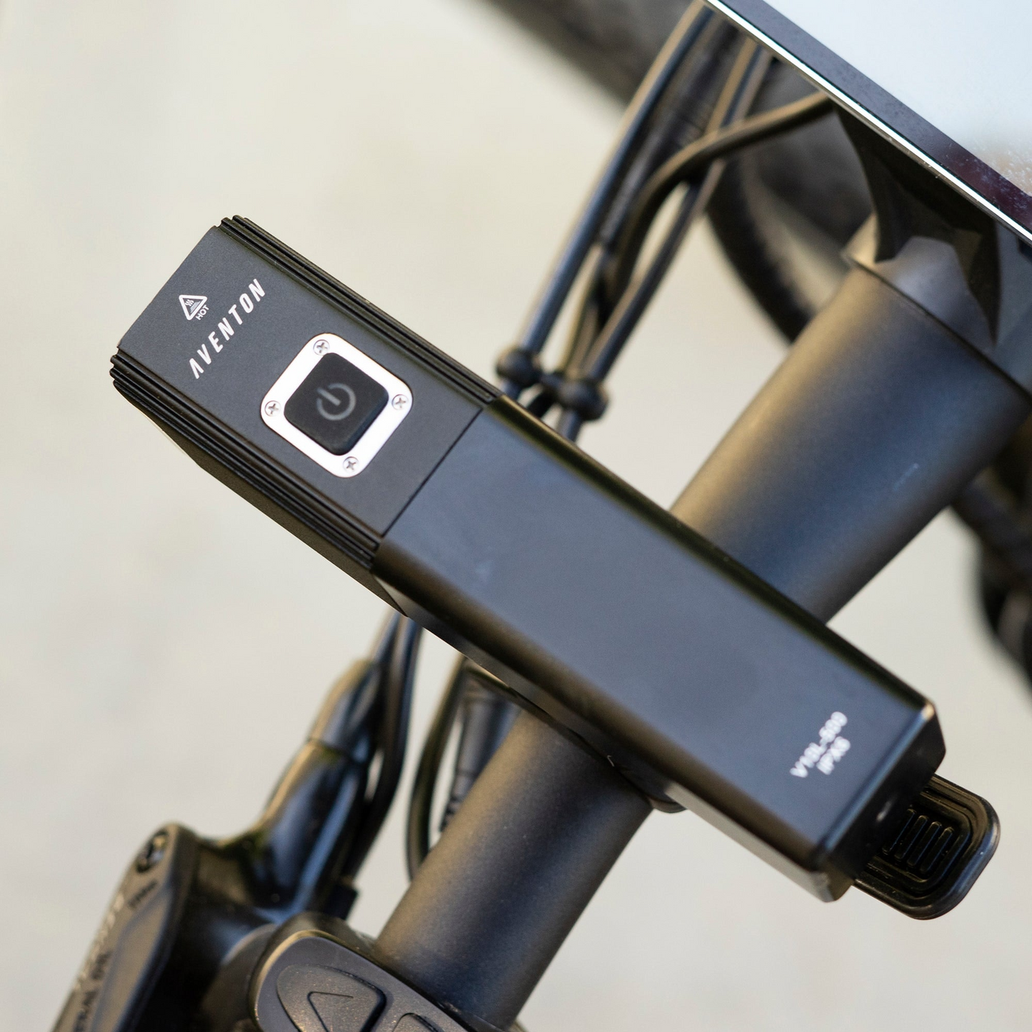 A close-up of a black Aventon V10-500 headlight mounted on handlebars, featuring a power button and labeled "BY-X4," highlights its USB rechargeable design for maximum bike compatibility.