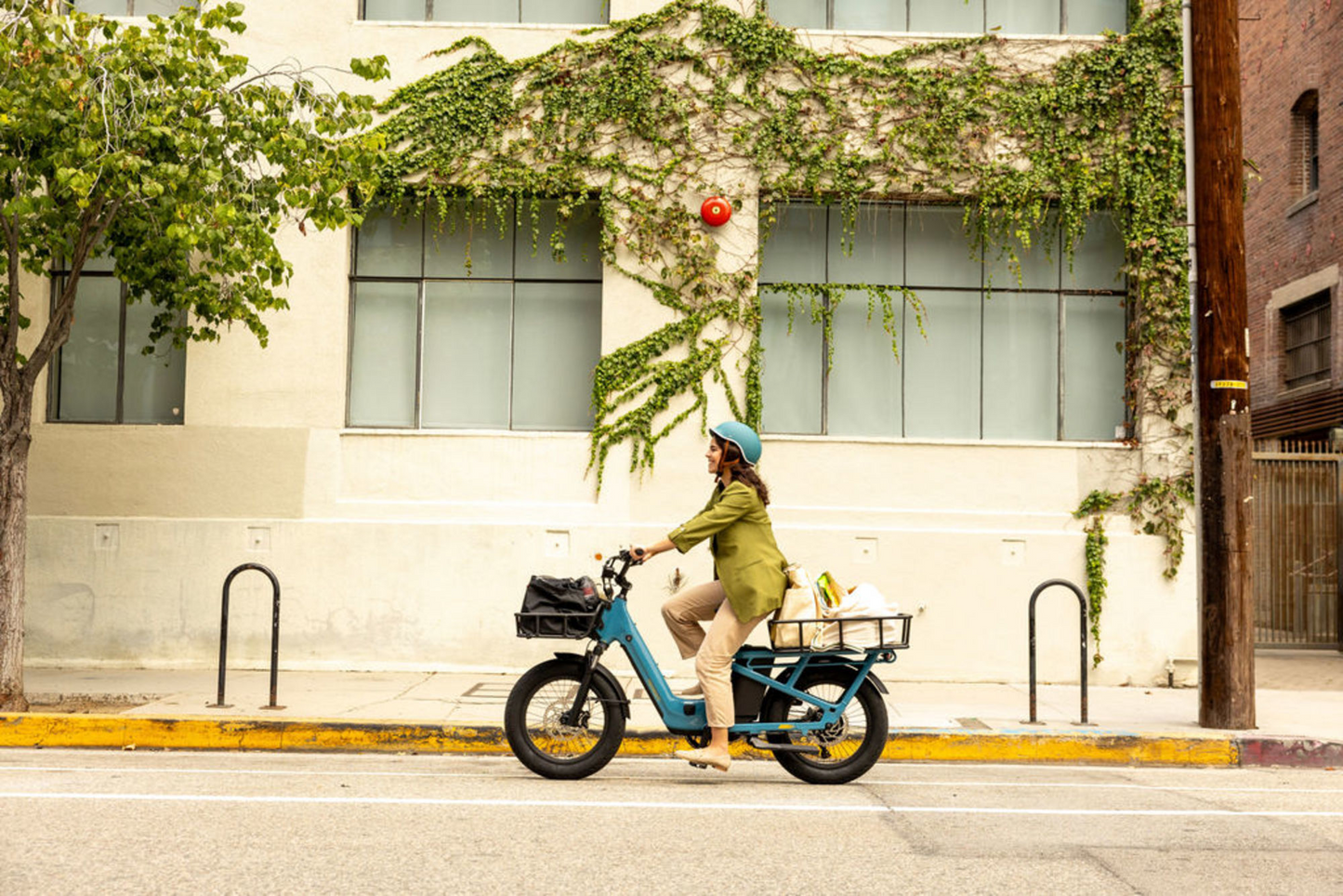 A person wearing a turban bikes past a building with ivy, carrying groceries in the bike's side baskets. They are riding a Momentum Cito E+ from Tampa Bay eBikes, enhancing their journey.