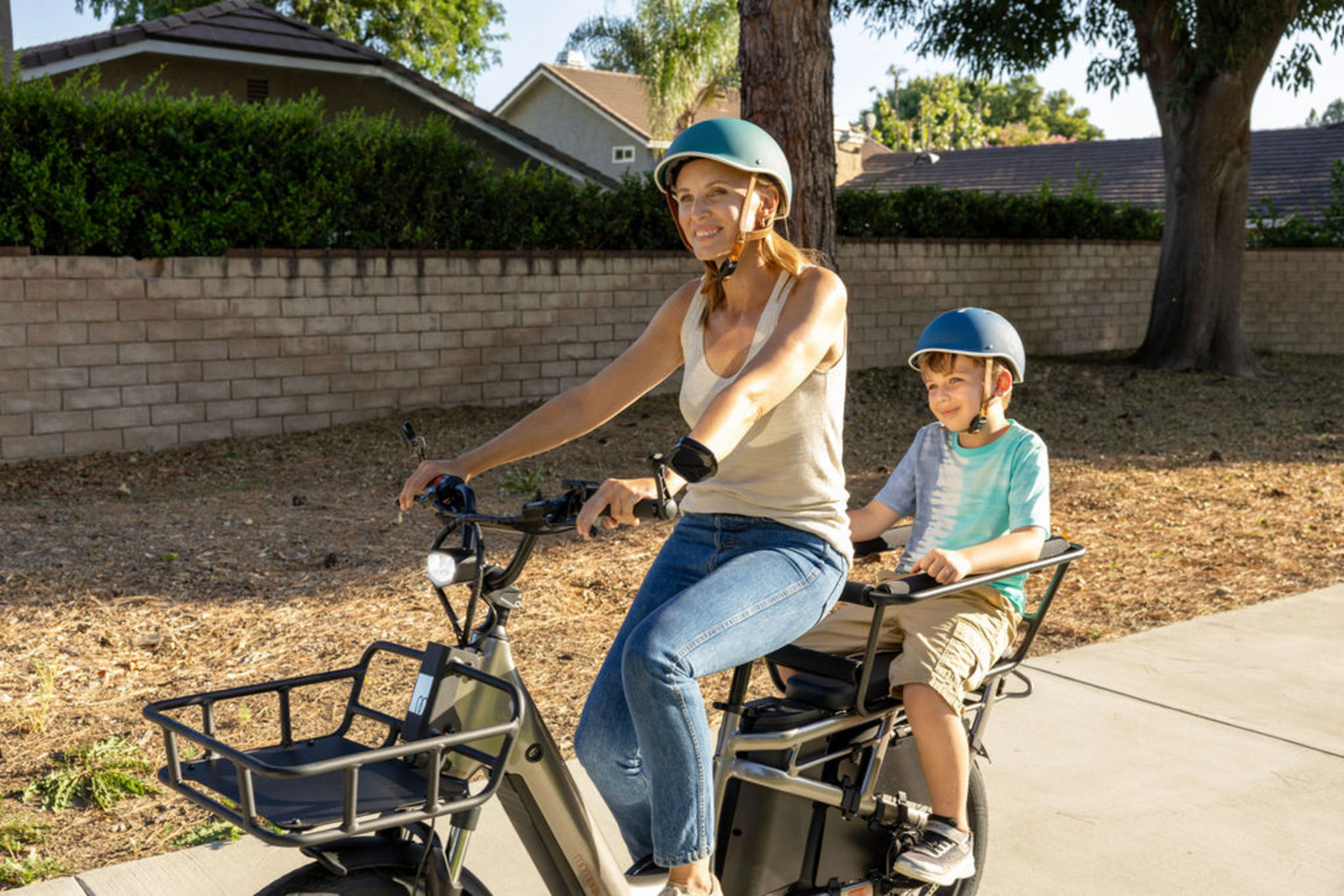 A woman and a young boy wearing helmets ride a Momentum - Cito E+ electric cargo bike by Tampa Bay eBikes along a suburban street.