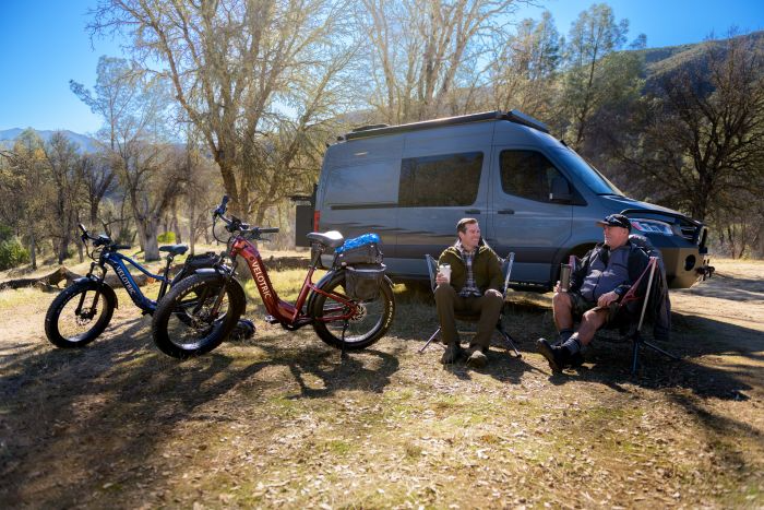 Two adventurers relax in camping chairs beside a parked van and their Velotric - Nomad 2 all-terrain eBikes, surrounded by trees and hills.