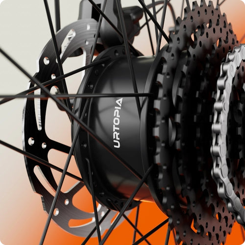 Close-up of the Urtopia - Carbon Classic eBike rear wheel hub with "URTOPIA" branding, highlighting spokes, gear cassette, disc brake rotor, and chain against a white and orange backdrop.