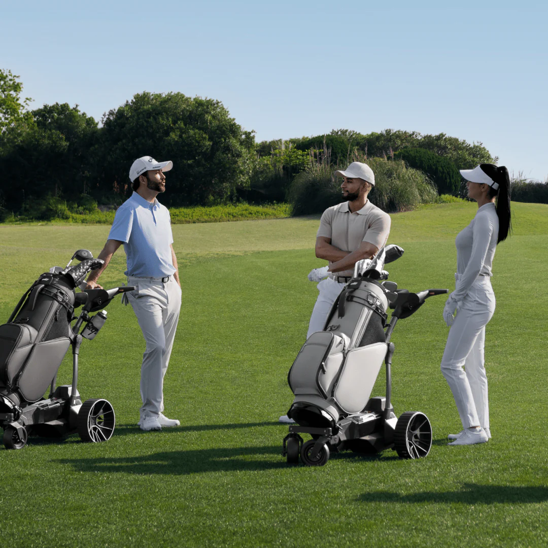 Three people in golf attire stand on a Tampa Bay course beside their Navee Birdie 3X electric push cart by Navee, chatting on a sunny day.