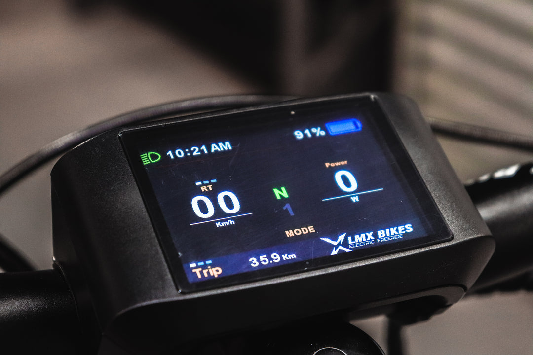 Close-up of an electric bike's digital display showing the time 10:21 AM, speed 0 km/h, power output 0, battery 91%, and trip distance 35.9 km. The display, from Tampa Bay eBikes, has the brand "LMX" at the bottom and a brushless mid-mounted motor providing smooth pedal assistance. The product is the LMX - 64 electric bike.