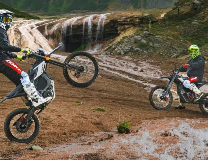 Two motorcyclists in helmets ride near a rocky waterfall on Segway Xaber 300 electric dirt bikes by Segway; one rider performs an off-road wheelie while the other stays still.