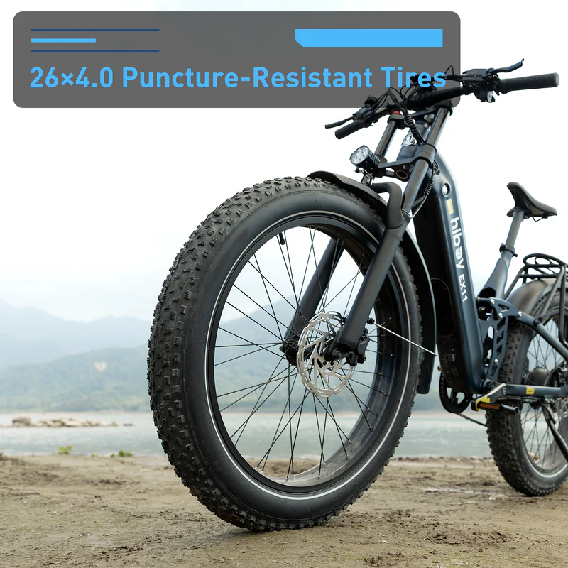 A close-up of the Hiboy - EX11, a black full suspension electric bike by Hiboy, featuring thick 26x4.0 puncture-resistant tires, parked on sand near water and mountains.