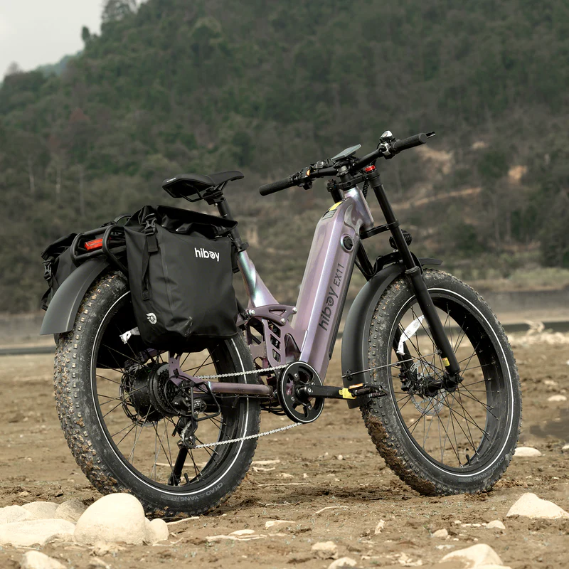 The Hiboy EX11 electric bike by Hiboy, featuring a 750W motor, fat tires, and pannier bags, is parked on rocky ground outdoors with a forested hillside in the background.