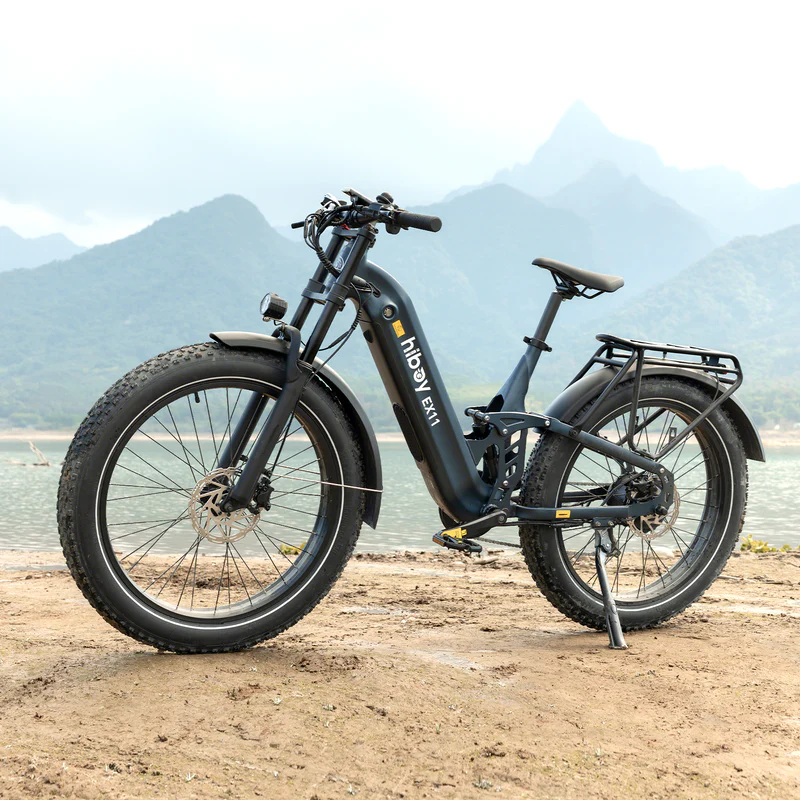 A black Hiboy - EX11 full suspension electric bike by Hiboy, equipped with a 750W motor, is parked on a dirt path beside a lake with mountains in the background.