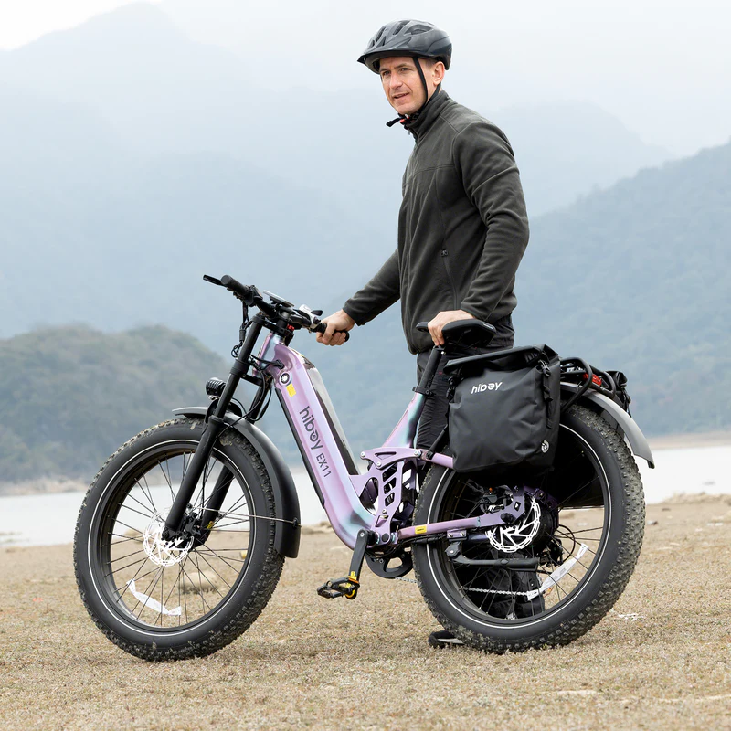 A man in a helmet and jacket stands by a body of water with mountains behind him next to a purple Hiboy - EX11 electric bike featuring 750W motor, fat tires, and a large black pannier bag.