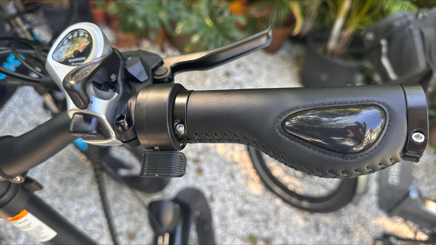 Close-up of a bicycle handlebar fitted with the ElectricAllWheel Thumb Throttle Conversion Kit (Lectric/AIPAS Fit), shown with visible stitching on the ergonomic grip, gear shifter, brake lever, and universal thumb throttle. Plants and gravel in back.