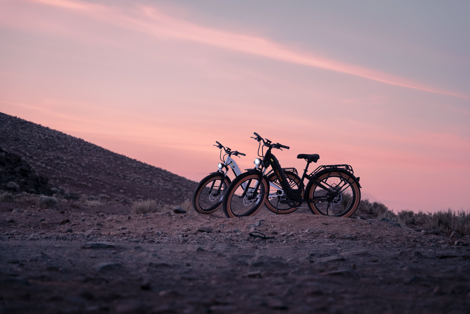 Two AIMA - Big Sur bicycles are parked on a rocky terrain during a pink and purple sunset.