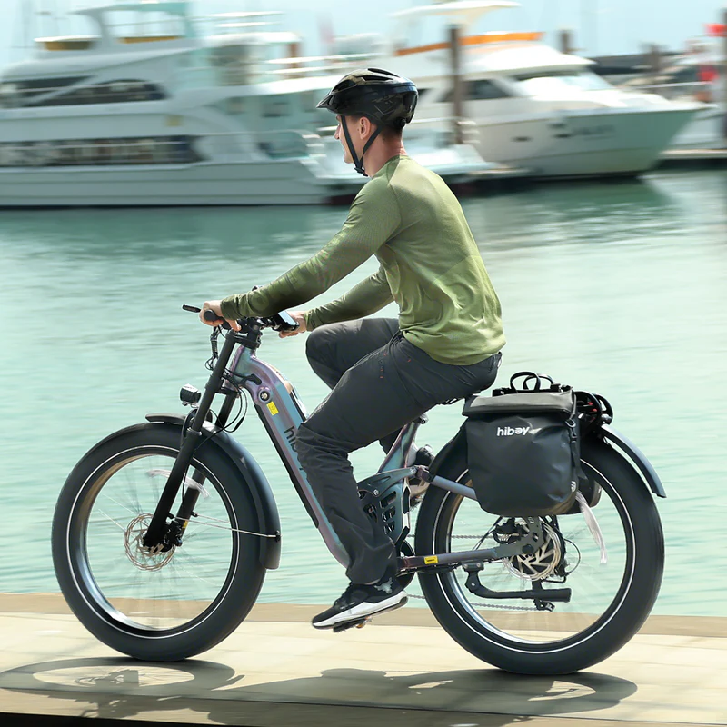 A person in a helmet and green long-sleeve shirt rides the Hiboy - EX11 commuter electric bike with a pannier bag along a waterfront, with boats visible in the background.