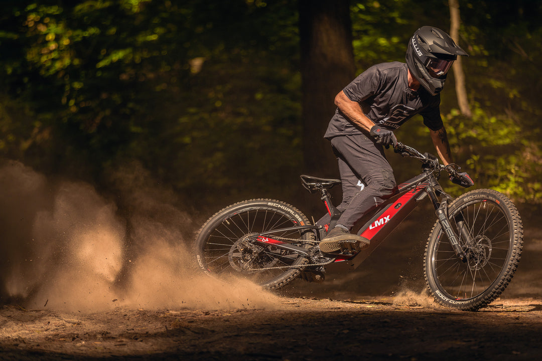 A mountain biker wearing a helmet and protective gear rides an LMX - 64 with LMX branding, kicking up dirt as they navigate a woodland trail.