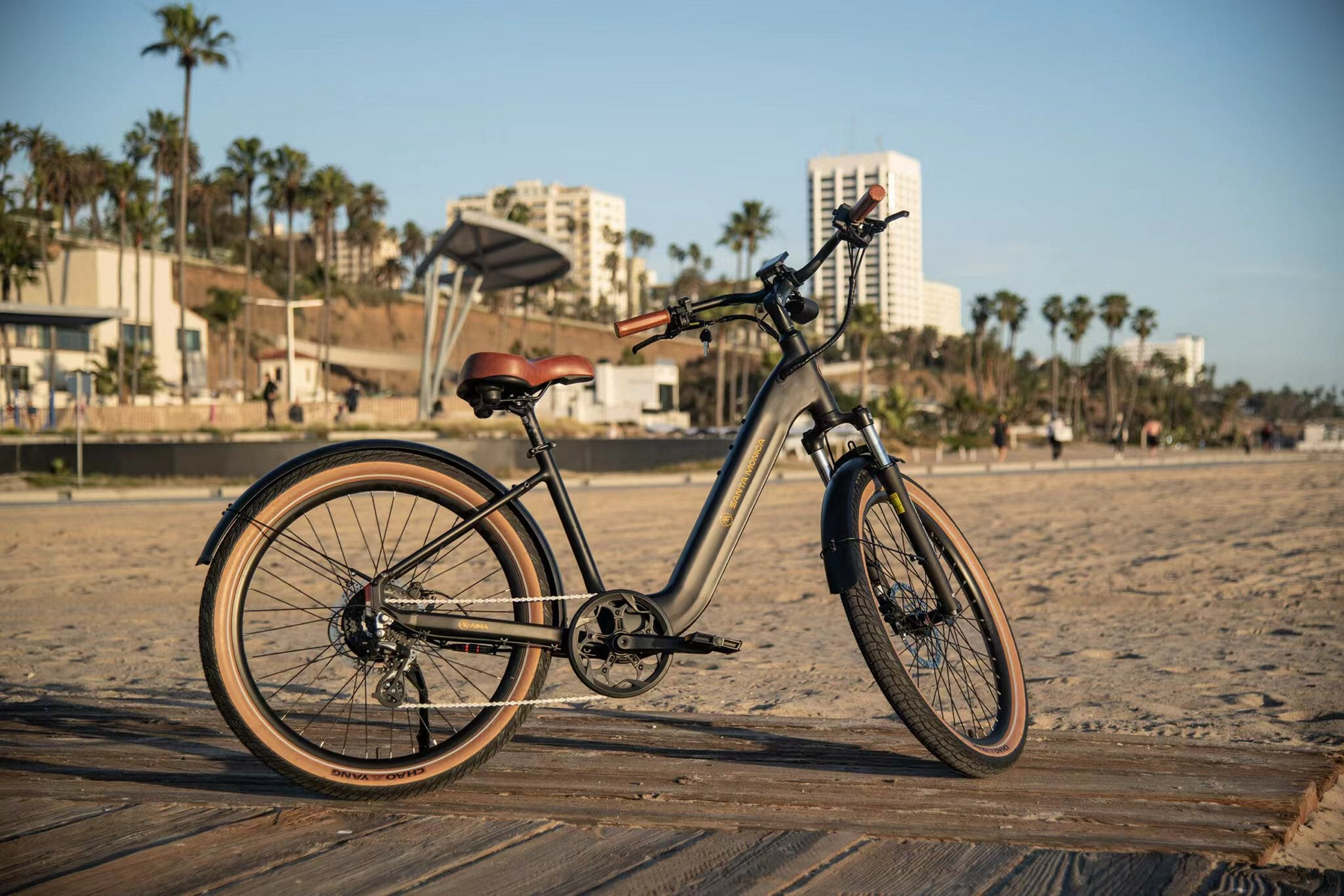 A modern electric bicycle equipped with a powerful 750W Bafang Motor is parked on a wooden boardwalk at the beach, perfect for urban commuting. The AIMA - Santa Monica by AIMA stands out against the backdrop as palm trees sway gently with the wind, and the iconic Santa Monica buildings loom proudly in the background.