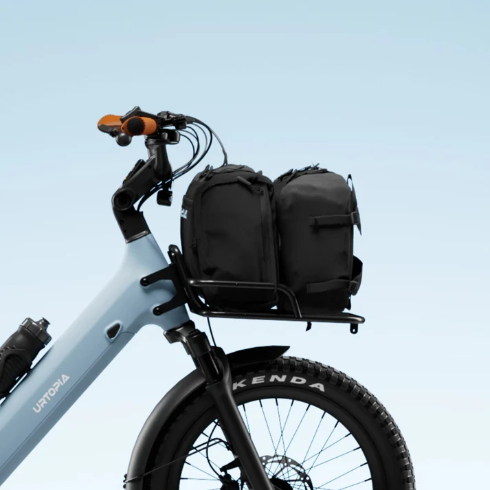 A Urtopia Joy Carbon Front Rack is attached to a light blue Urtopia bicycle, carrying two black bags, all set against a plain light blue background.