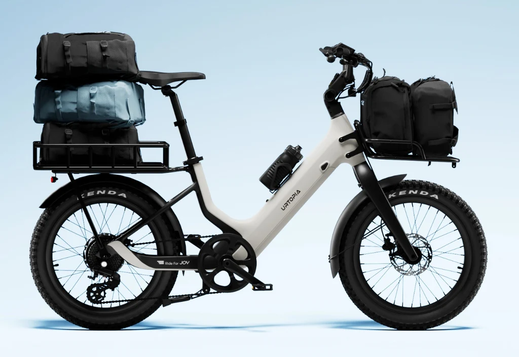 The Urtopia Carbon Joy Ultra electric cruiser cargo bike by Urtopia, featuring large tires and loaded with bags on both racks, is displayed against a plain light blue background.