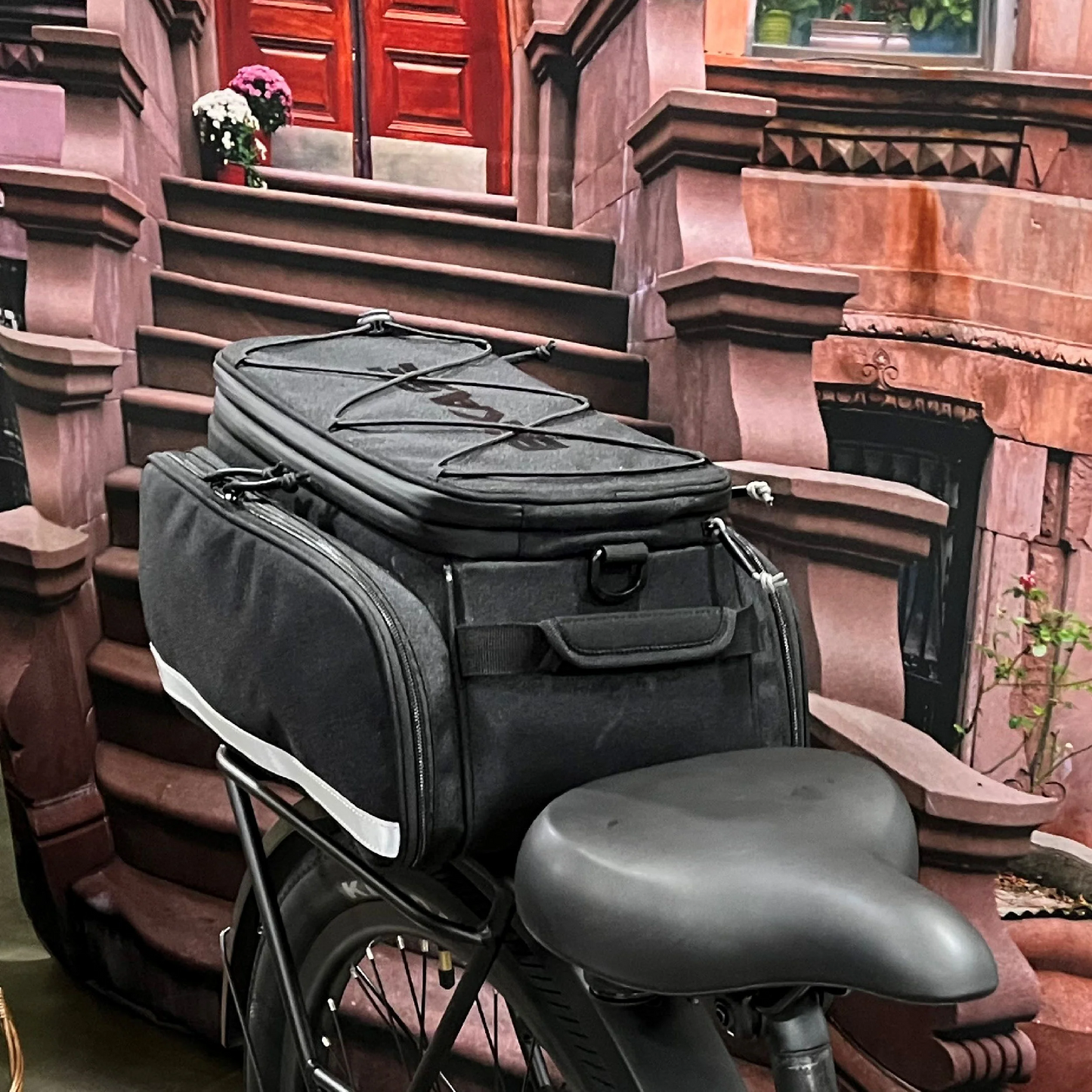 The Bikase Bag - Big Poppa MIK, featuring expandable panniers, is displayed mounted on a bike in front of a brick building and stairs.