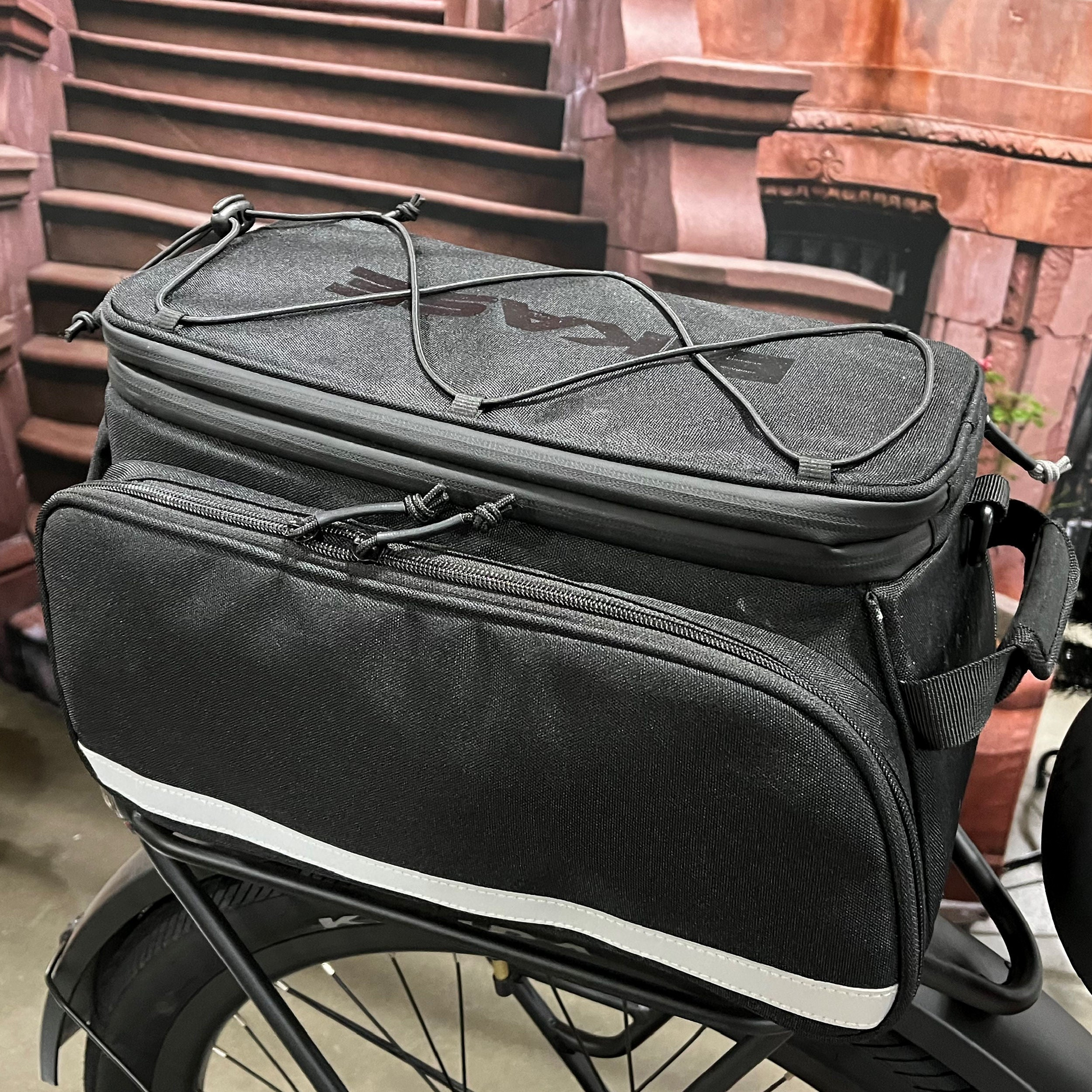 The black Bikase Big Poppa MIK bicycle pannier, with expandable compartments and waterproof features, securely attaches to a rear bike rack. It includes zippered sections and bungee cords on top, set against a backdrop of brick stairs.