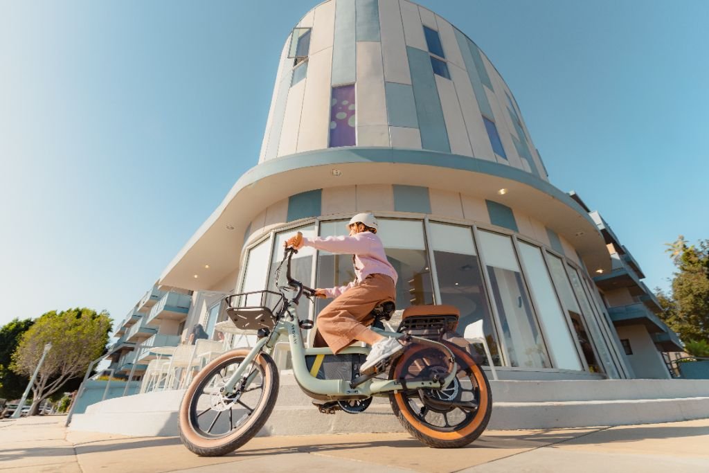 A person rides a Segway Muxi electric utility bike by Segway, with an 80-mile range, along a sidewalk in front of a modern curved building under clear skies.