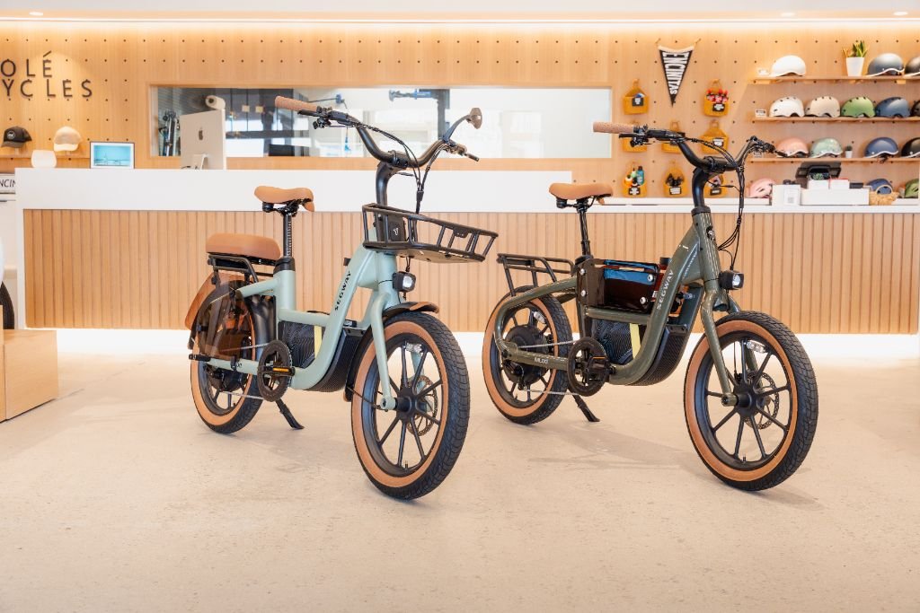 Two Segway Muxi electric utility bikes with tan seats and tires are displayed indoors at a modern retail counter lined with helmets and accessories, highlighting their impressive 80-mile range.