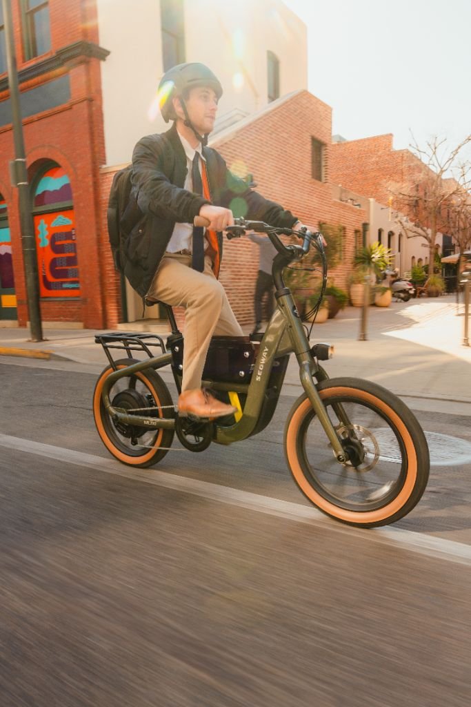 A man in business attire and a helmet rides a Segway Muxi electric utility bike from Segway along a city street, with brick buildings in the background.