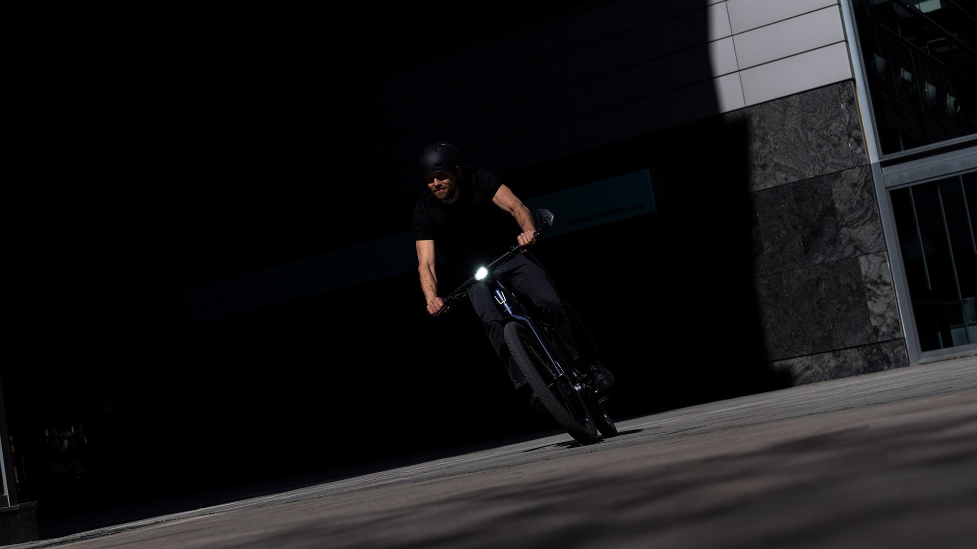 A man in black clothing and helmet rides an eBike, the Stromer ST7 Alinghi Red Bull Racing Edition, in front of a modern building in a shaded urban setting.