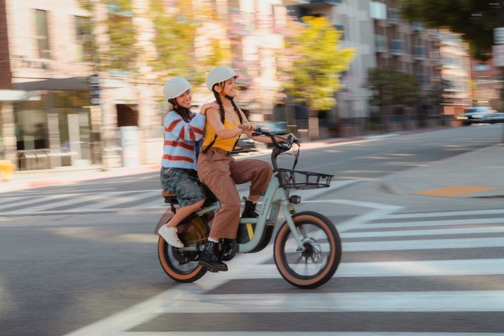 Two people in helmets ride a Segway Muxi by Segway, featuring a 750W motor and 80-mile range, together through a sunny city street crosswalk.