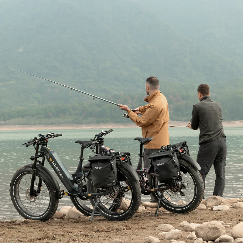Two men fish by a lake with two Hiboy EX11 electric bikes from Hiboy, each featuring a 750W motor and full suspension, parked on rocky ground; mountains and water appear in the background.