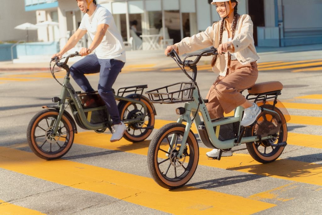 Two people wearing helmets and casual clothes ride Segway Muxi electric bikes with powerful 750W motors, crossing a sunny urban street marked with yellow lines.
