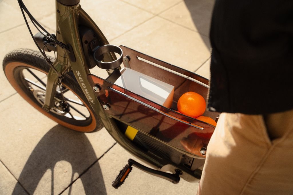 A close-up of a Segway Muxi by Segway shows its open storage compartment holding a cup, a white box, and an orange on a sunny day.
