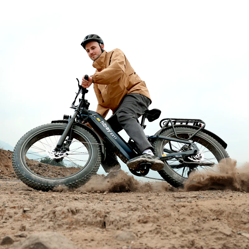 A man in a helmet and tan jacket rides the Hiboy - EX11, a black 750W fat tire electric bike by Hiboy, across sandy, rugged terrain, kicking up dirt behind him.