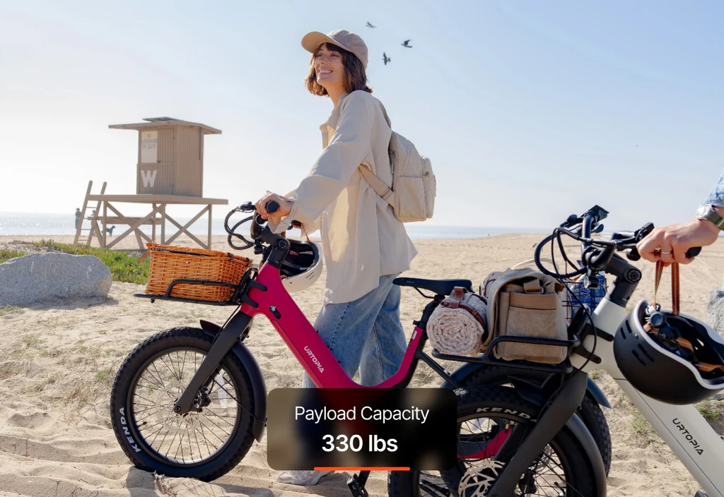 On a sandy beach, a woman holds the Urtopia - Carbon Joy Ultra e-bike with basket. Text overlay: "Payload Capacity 330 lbs." Another bike and a beach hut appear in the background. Brand: Urtopia.
