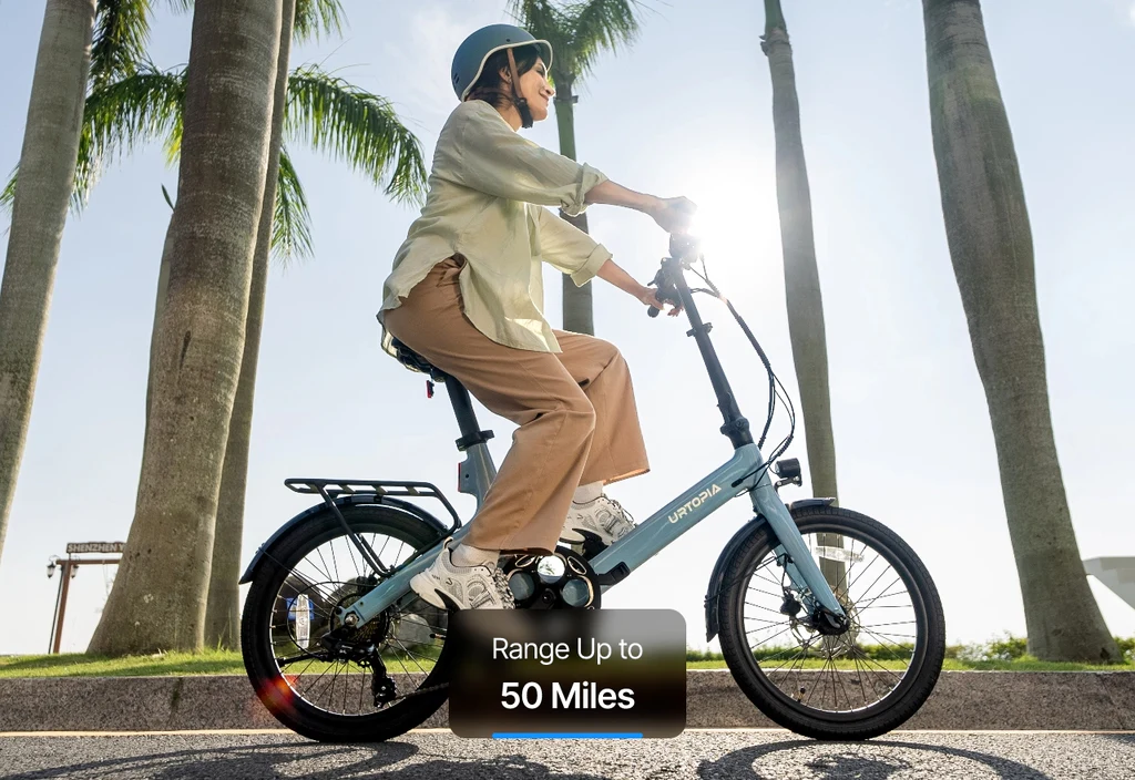 A woman in a helmet rides the Urtopia - Carbon Fold Step-Thru by Urtopia, a blue lightweight electric bike, along a palm-lined road; a label highlights its range of up to 50 miles.