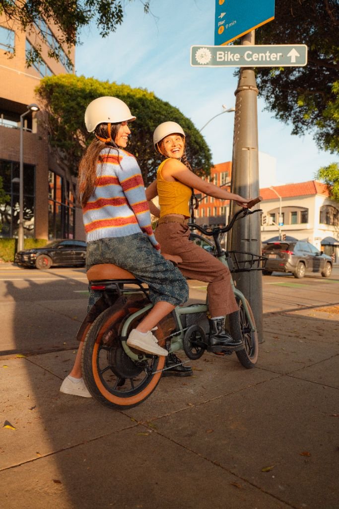 Two women in helmets sit on a Segway Muxi electric utility bike by a "Bike Center" sign at a street corner, enjoying their Segway ride with up to 80 miles of range on a sunny day.