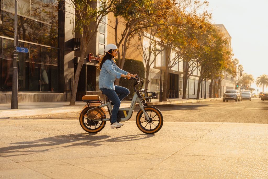 A person in a helmet and denim jacket rides a Segway Muxi electric bike from Segway along a sunlit city street lined with trees and parked cars.