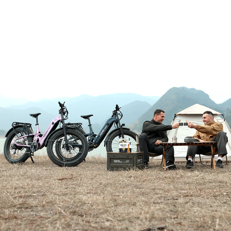 Two people toast drinks at a campsite table, with two Hiboy EX11 full suspension electric bikes parked nearby and mountains in the background.