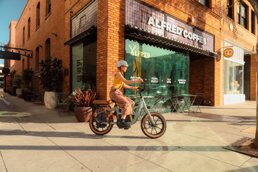 A helmeted rider cruises past Alfred Coffee on a sunny city street lined with brick buildings, riding a Segway Muxi electric utility bike by Segway with an 80-mile range.