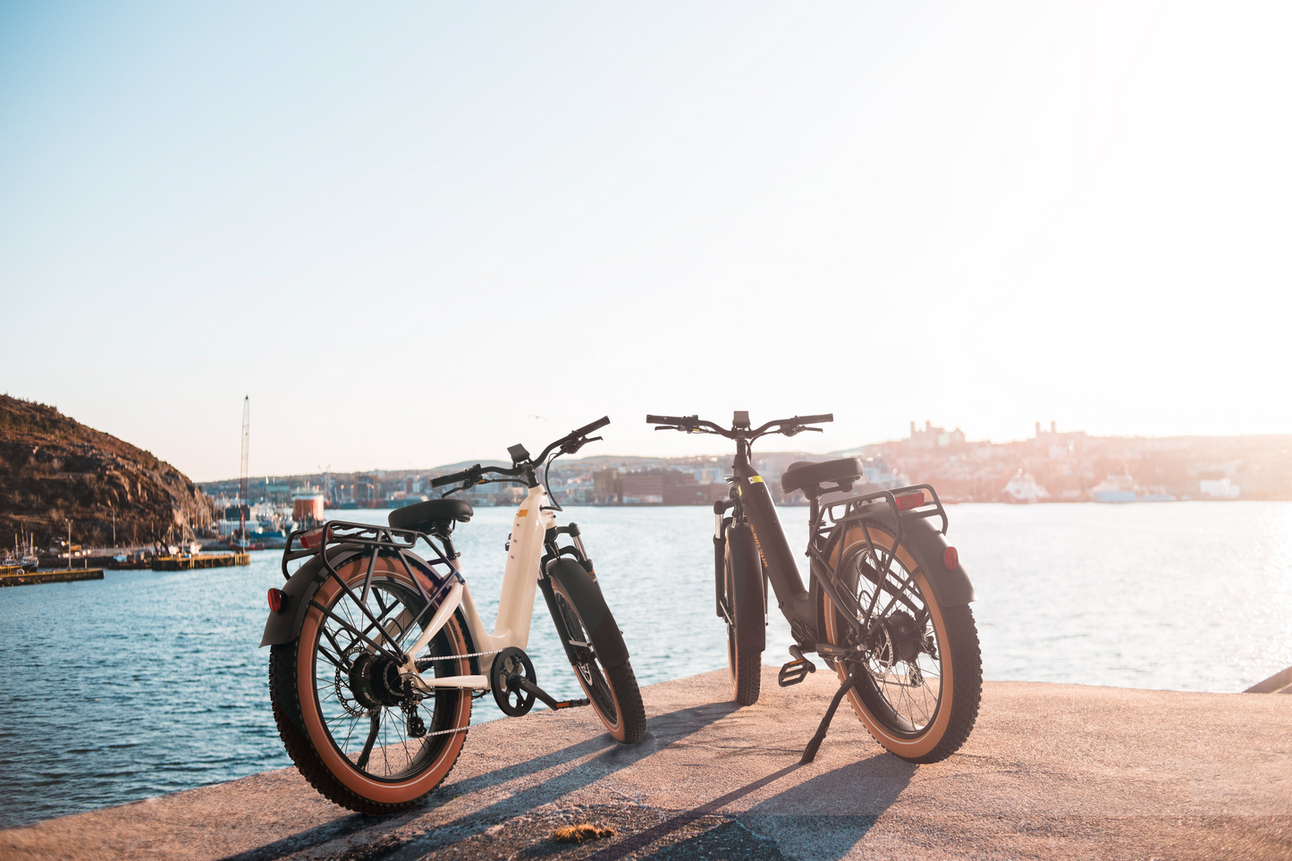 Two AIMA - Big Sur eBikes are parked on a waterfront, overlooking a body of water with ships in the distance under a clear sky. Equipped with powerful motors, these all-terrain bikes from AIMA promise an exciting ride along the scenic shoreline.