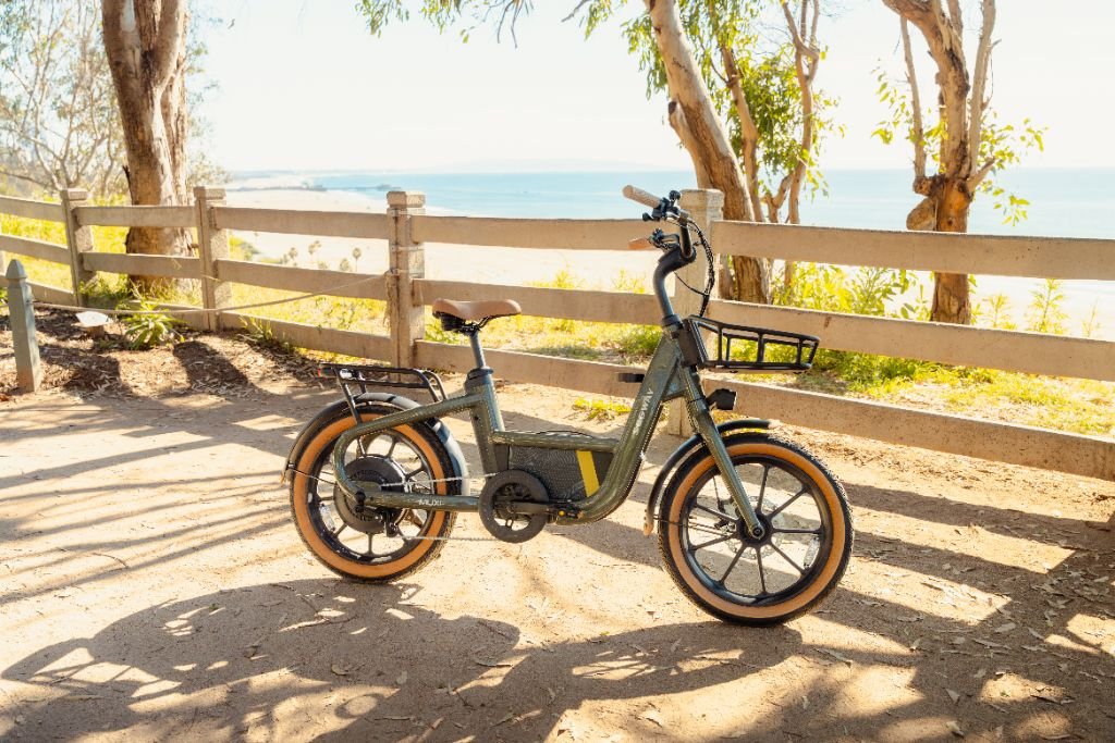 A Segway Muxi electric utility bike with a front basket is parked on a sunny dirt path by a wooden fence, overlooking the beach and ocean—a great place to enjoy the impressive range of this Segway eBike.