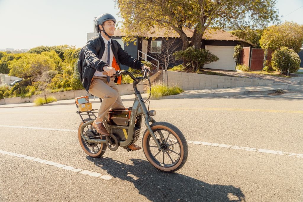 A helmeted rider with a backpack cruises uphill on a sunny day, passing houses and trees, aboard the Segway Muxi electric utility bike by Segway.