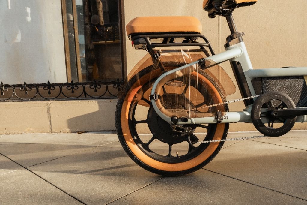 Close-up of the rear wheel, seat, and frame of a Segway Muxi electric utility bike by Segway with tan tires, parked on a sidewalk beside a beige wall and window.