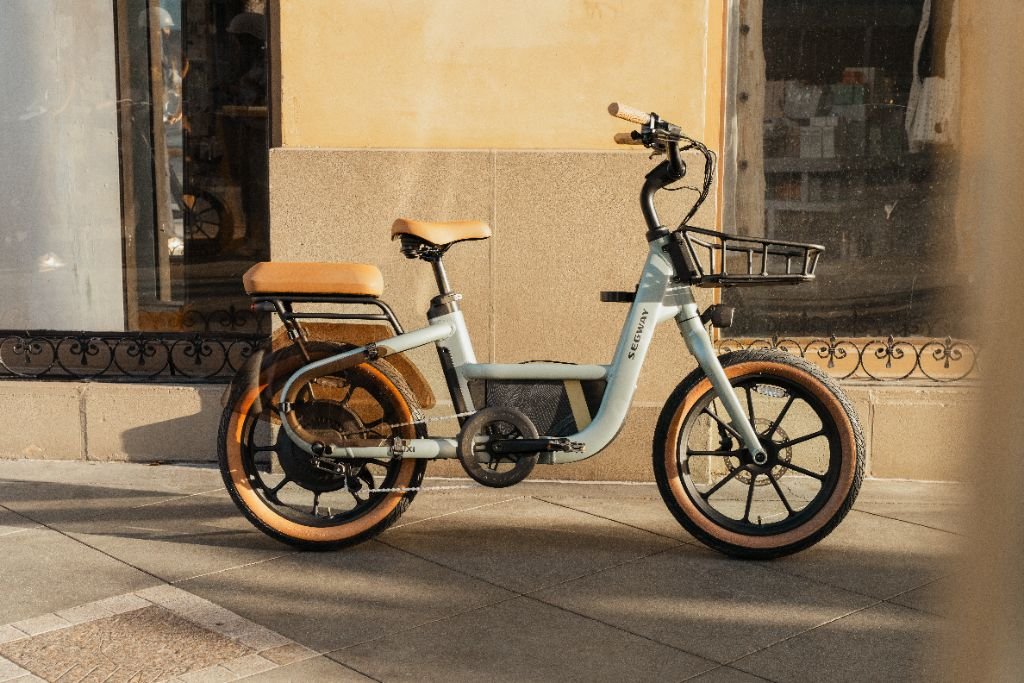 A Segway Muxi electric utility bike in light blue with a tan seat and tires is parked on a sidewalk outside a building with large windows.