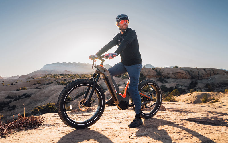 A man in a helmet stands with a HeyBike - Hero (Mid-Drive) on rocky terrain under a clear sky.