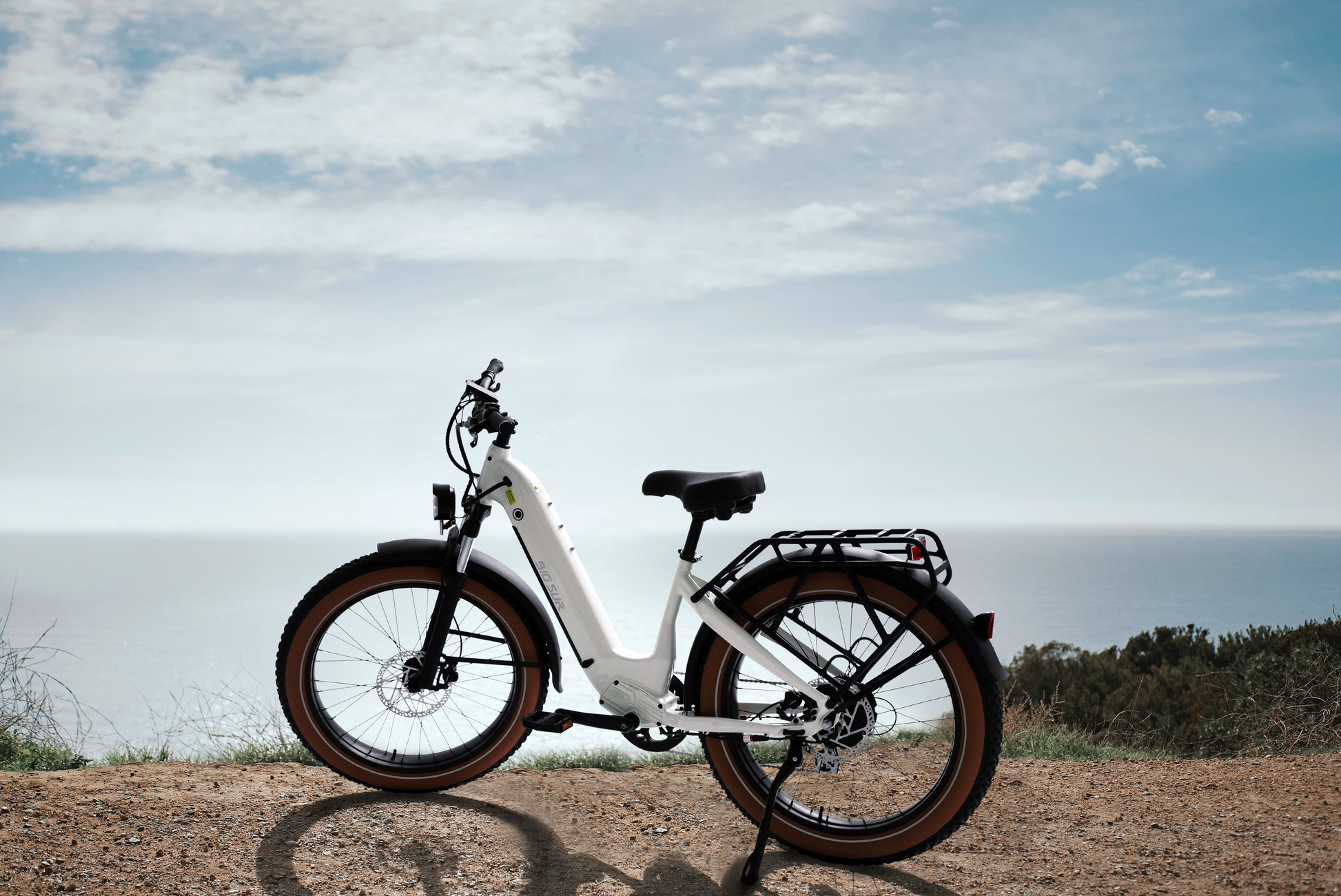 An AIMA - Big Sur eBike, featuring a powerful motor and brown tires, is parked on a dirt path, overlooking the ocean beneath a partly cloudy sky.