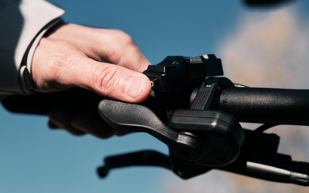 A close-up shows a hand pressing a button on the Velotric Discover M handlebar with gear and brake controls against a clear blue sky, highlighting this comfort commuter eBike from Velotric.