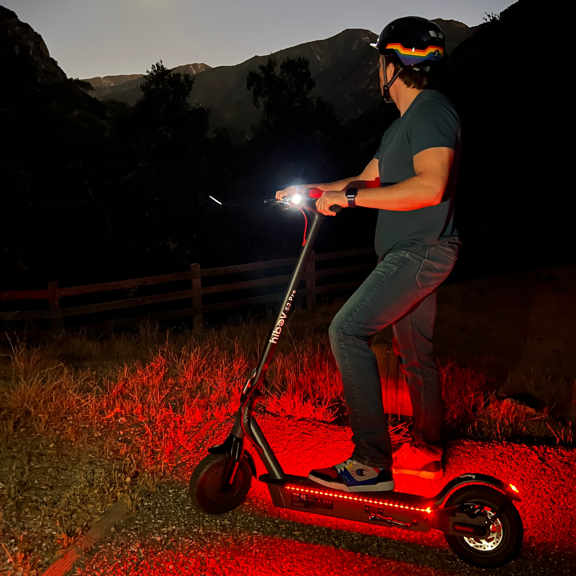 A person wearing a helmet rides a Hiboy - S2 Pro Electric Scooter with red lights at dusk, effortlessly maneuvering through the mountains and trees in the background, ideal for high-performance daily commuting.